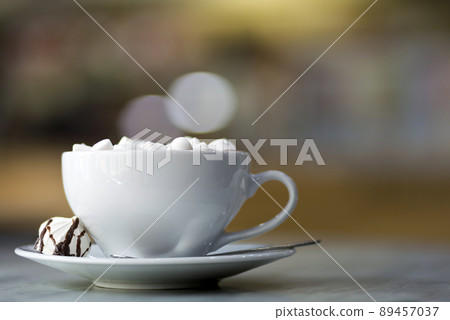 Mug of coffee with marshmallows on porcelain plate on blurred colorful interior bokeh background. Mug of coffee with marshmallows on porcelain plate on blurred colorful interior bokeh background. 89457037