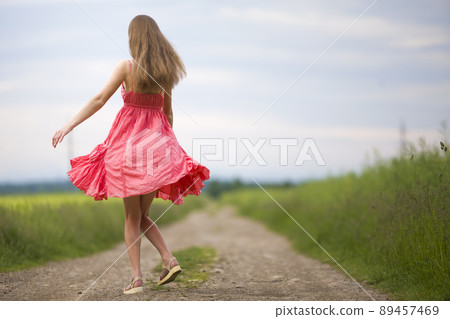 Back view of young romantic slim woman in red dress with long hair walking by ground road along green field on sunny summer day on blue sky copy space background. Back view of young romantic slim woman in red dress with long hair walking by ground road along green field on sunny summer day on blue sky copy space background. 89457469