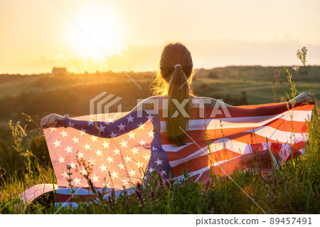 Back view of happy woman with USA national flag sitting outdoors at sunset. Positive girl celebrating United States independence day. International day of democracy concept. Back view of happy woman with USA national flag sitting outdoors at sunset. Positive girl celebrating United States independence day. International day of democracy concept. 89457491