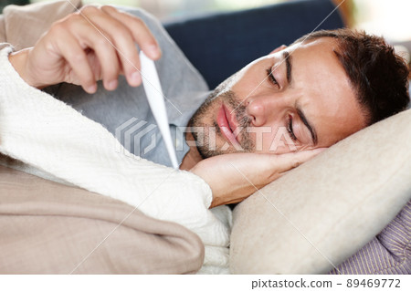 Keeping a close eye on his temperature. Shot of a young man lying on the sofa reading his temperature on a thermometer. 89469772