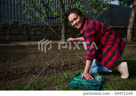 Charming woman gardener using garden tools loosens and digs black soil near a flower bed with growing greenery in a country garden 89472284