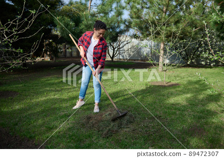 Full length of female gardener raking grass clippings into a compost heap in her garden of a countryside house backyard Full length of female gardener raking grass clippings into a compost heap in her garden of a countryside house backyard 89472307