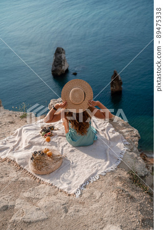 Street photo of a beautiful woman with dark hair in a mint dress and hat having a picnic on a hill overlooking the sea. Street photo of a beautiful woman with dark hair in a mint dress and hat having a picnic on a hill overlooking the sea. 89475338