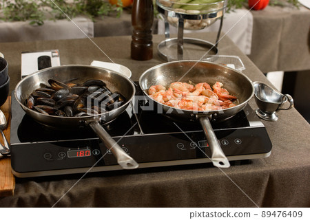 two pans with mussels and shrimp on an induction stove two pans with mussels and shrimp on an induction stove 89476409