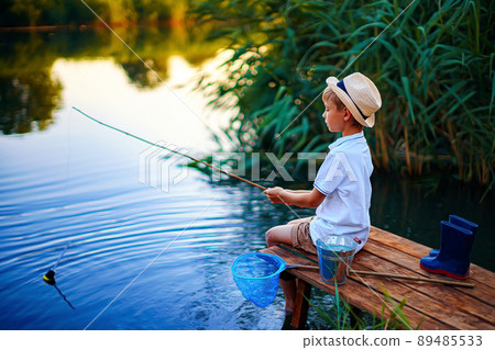 Boy in straw hat with a fishing rod catches fish on riverbank at golden hour of sunset, river 89485533