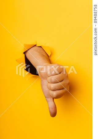 Hand showing a thumb down through ripped hole in yellow paper background. Concept of dislike and disapproval gesture. Vertical image 89488758
