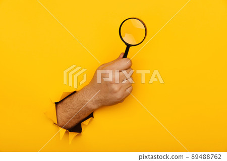 Hand holds black magnifying glass in torn hole of yellow background. Searching information concept Hand holds black magnifying glass in torn hole of yellow background. Searching information concept 89488762