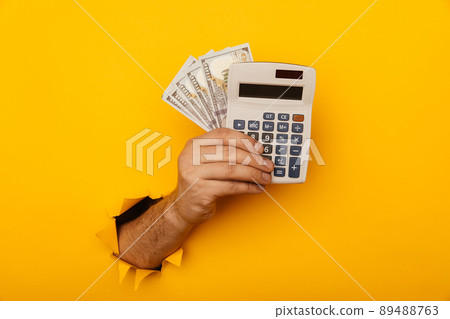 Male hand with calculator and dollar banknotes through a paper hole in yellow background 89488763