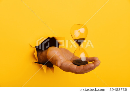 Male hand holds hourglass through torn hole yellow paper Male hand holds hourglass through torn hole yellow paper 89488786