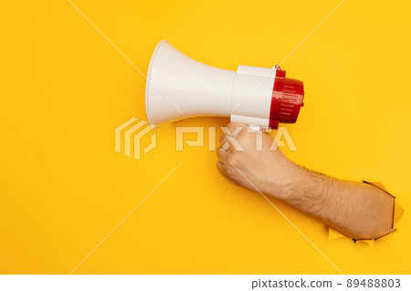 Hand holds a megaphone from a hole in the wall on a yellow background with copy space 89488803