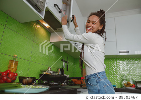 Young mixed-race woman preparing food in kitchen, happy casual housewife opens cupboard Young mixed-race woman preparing food in kitchen, happy casual housewife opens cupboard 89503467
