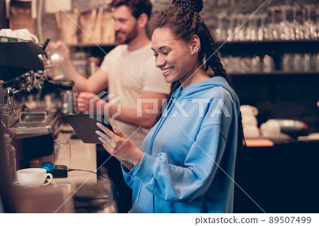 Smiled stylish young woman barista smiling and using tablet device. Learning coffee making. 89507499