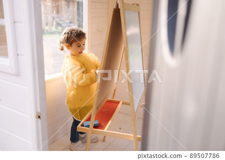Young artist paints on wooden boards with chalk. Cute girl in yeallow raincoat Young artist paints on wooden boards with chalk. Cute girl in yeallow raincoat 89507786
