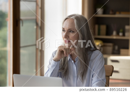 Pensive dreamy older 60s business lady sitting at laptop computer 89510226