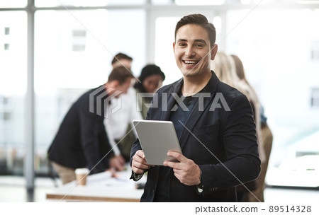 Technology adds so much ease into the workplace. Portrait of a young businessman using a digital tablet in an office with his colleagues in the background. Technology adds so much ease into the workplace. Portrait of a young businessman using a digital tablet in an office with his colleagues in the background. 89514328
