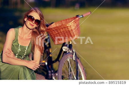 Perfect day for a bike ride in the countryside. Shot of a beautiful young woman relaxing in the park next to her old fashioned bicycle. 89515819