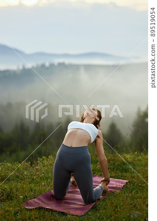 Front view of pretty female wearing white top and grey leggings bending back touching legs with arms outdoors. Fit brunette girl holding camel pose among grass in morning nature. Concept of yoga. 89519054