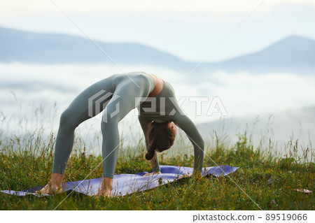 Side view of brunette female doing urdhva dhanurasana practice among grass in morning nature. Young fit girl exercising alone standing in wheel pose outdoors. Concept of yoga and healthy lifestyle. 89519066