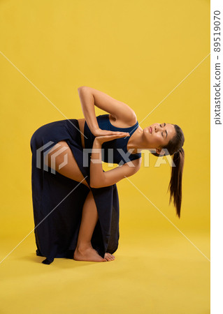 Front view of flexible girl practicing yoga pose indoors. Brunette female standing with bent legs, bending forward, with prayer hands, looking up. Concept of new age and yoga. Front view of flexible girl practicing yoga pose indoors. Brunette female standing with bent legs, bending forward, with prayer hands, looking up. Concept of new age and yoga. 89519070