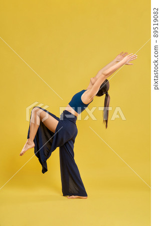 Side view of slim woman standing alone, raising leg and hands. Flexible brunette female with pony tail stretching, smiling, yoga doing. Isolated on yellow studio background. Side view of slim woman standing alone, raising leg and hands. Flexible brunette female with pony tail stretching, smiling, yoga doing. Isolated on yellow studio background. 89519082