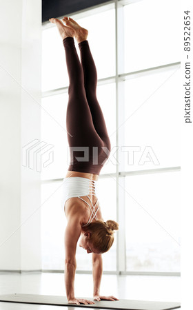 Young slim woman in leggings doing handstand with straight legs in modern yoga studio Young slim woman in leggings doing handstand with straight legs in modern yoga studio 89522654