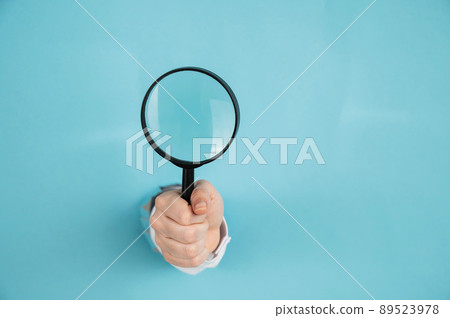 A woman's hand sticking out of a hole in a paper blue background holds a magnifying loupe. Copy space.  89523978