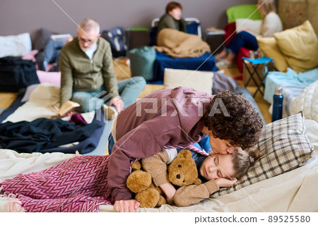 Young woman in casualwear kissing her cute little son sleeping with teddybear Young woman in casualwear kissing her cute little son sleeping with teddybear 89525580