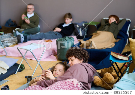 Young female refugee and her son with smartphone lying under blanket Young female refugee and her son with smartphone lying under blanket 89525597