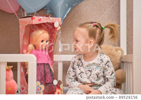 A little girl in pajamas receives birthday gifts while sitting in a white baby cot. Balloons in the background. A little girl in pajamas receives birthday gifts while sitting in a white baby cot. Balloons in the background. 89528156