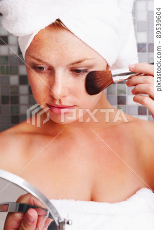 Cute woman looking at self at hand mirror and applying make up. Lovely young female looking at self at hand mirror and applying make up. 89539604