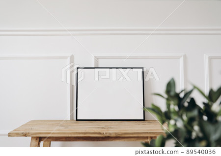 Blank horizontal black picture frame mockup. Old wooden console table. White wall moulding background, trim decor. Elegant home interior, still life photo. Art dispaly. Blurred green leaves, plant 89540036