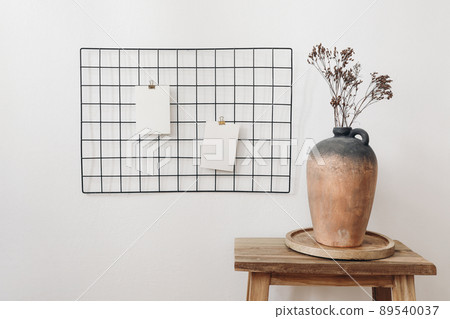 Rustic clay vase with dry hypericum bouquet on wooden table. Black metal mesh noticeboard, bulletin board with blank memo cards mockups. Elegant home office interior concept. White wall background. 89540037