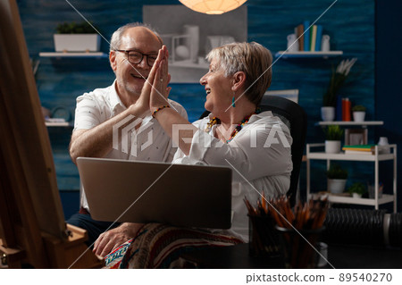 Elderly artist couple holding laptop doing high five hand gesture celebrating selling artwork in online auction sitting in home studio. Senior art creators enjoying success on social media. Elderly artist couple holding laptop doing high five hand gesture celebrating selling artwork in online auction sitting in home studio. Senior art creators enjoying success on social media. 89540270