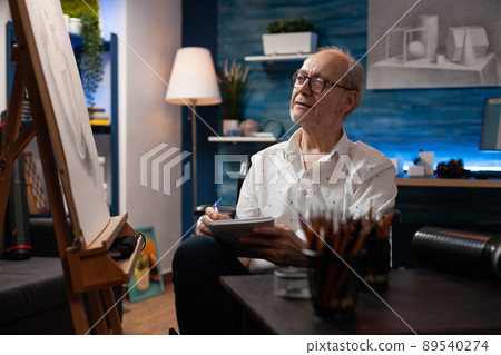 Senior hobby art creator taking notes keeping journal of learning progress in creative workshop looking at easel in home art studio. Elderly man artist sitting in wheelchair doing sketch on notebook. 89540274