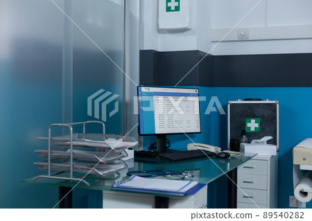 Empty doctor office with nobody in it equiped with medical equipment having work computer on desk and first aid kit on wall. Modern hospital room with rollboxes and clipboard cases reports. 89540282