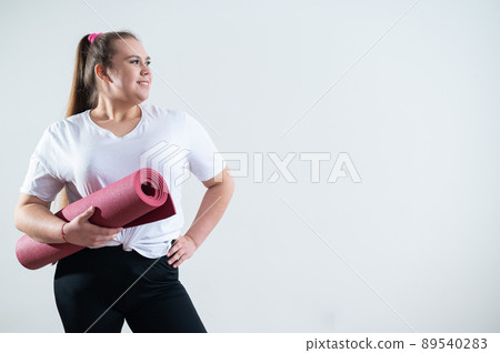 Young fat caucasian woman holding a sport mat. Charming plus size model in sportswear stands on a white background Young fat caucasian woman holding a sport mat. Charming plus size model in sportswear stands on a white background 89540283
