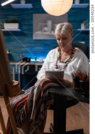 Elderly artist doing sketch on notebook for practice before drawing on easel in home art studio. Senior hobby art creator taking notes keeping journal of learning progress in workshop. 89540284