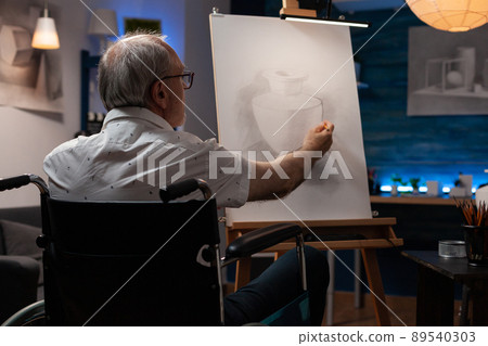 Older man who is paralyzed shading sketch in light study in evening class feeling inspired drawing vase model on paper. Focused retired art teacher using wheelchair doing creative artwork in pencil. Older man who is paralyzed shading sketch in light study in evening class feeling inspired drawing vase model on paper. Focused retired art teacher using wheelchair doing creative artwork in pencil. 89540303