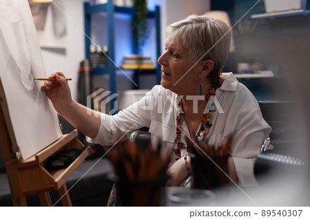 Elderly artist shading static nature model of vase using pencil on paper canvas in home creative workshop. Senior woman feeling creative drawing charcoal sketch on easel in art studio. Elderly artist shading static nature model of vase using pencil on paper canvas in home creative workshop. Senior woman feeling creative drawing charcoal sketch on easel in art studio. 89540307