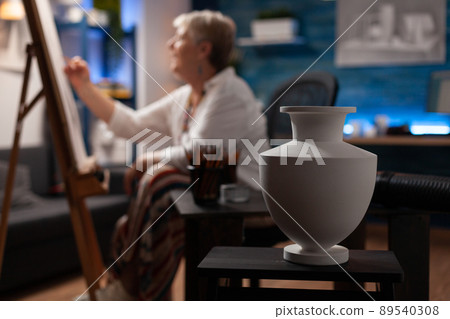 Closeup of white vase on wooden table used for inspiration by elderly artist sketching on easel in creative art studio. Selective focus on still life used as subject for drawing in workshop. 89540308