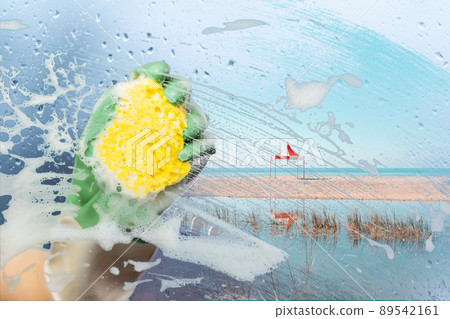 A hand in a rubber glove wipes the window glass with a sponge with foam and washes away dirt and bad weather. Outside the window is summer and the sea beach. Tourism and travel concept A hand in a rubber glove wipes the window glass with a sponge with foam and washes away dirt and bad weather. Outside the window is summer and the sea beach. Tourism and travel concept 89542161