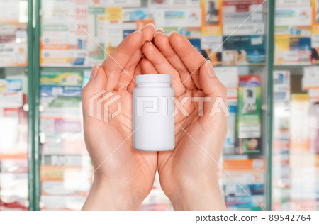 Close-up of female's palms holding a white jar of medicine. In the background, blurred pharmacy windows. Mock up. Pharmaceutical business concept Close-up of female's palms holding a white jar of medicine. In the background, blurred pharmacy windows. Mock up. Pharmaceutical business concept 89542764