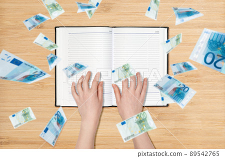 Female's hands lies on an empty book, euro banknotes are pouring down from above. Flat lay. The concept of the financial crisis and the economy Female's hands lies on an empty book, euro banknotes are pouring down from above. Flat lay. The concept of the financial crisis and the economy 89542765