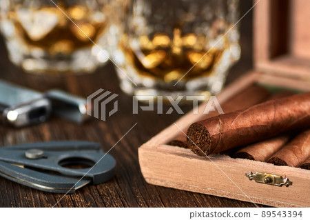 View of a wooden box with Cuban cigars, a lighter and a cutter. Still life with two glasses of whiskey. 89543394