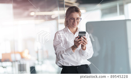 Businesswoman wearing glasses with mobile phone in hand standing in modern office 89545228