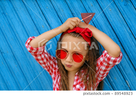 A girl with a red bow on her head and sunglasses holds a triangular shaped lollipop in hands. The child looks into the camera. Blue old painted Background from planks 89548923