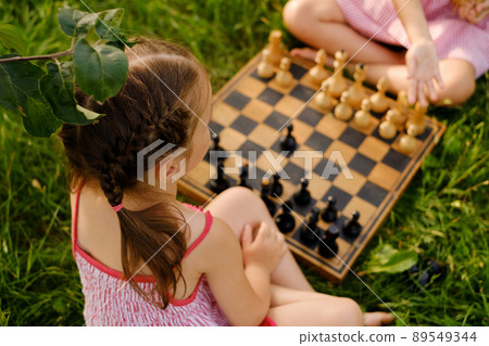 Two little girls are playing chess on a wooden chessboard outdoors in nature. One of the girls teaches the game and the rules of another child. Close-up. Rear view 89549344
