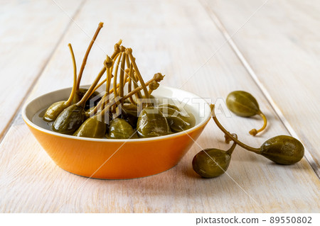Tasty pickled caper berries in an orange bowl on a white wooden table. Tasty pickled caper berries in an orange bowl on a white wooden table. 89550802