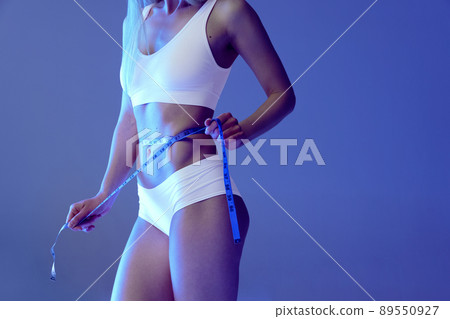 Cropped image of cropped slim tanned woman body in white underwear on blue studio background in neon. Measuring 89550927