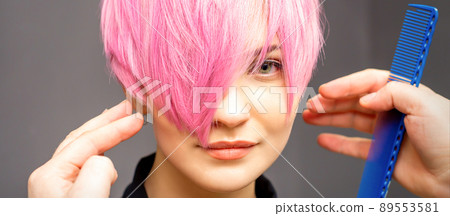 Hairdresser with hands and comb is checking out and fixing the short pink hairstyle of the young white woman in a hair salon Hairdresser with hands and comb is checking out and fixing the short pink hairstyle of the young white woman in a hair salon 89553581